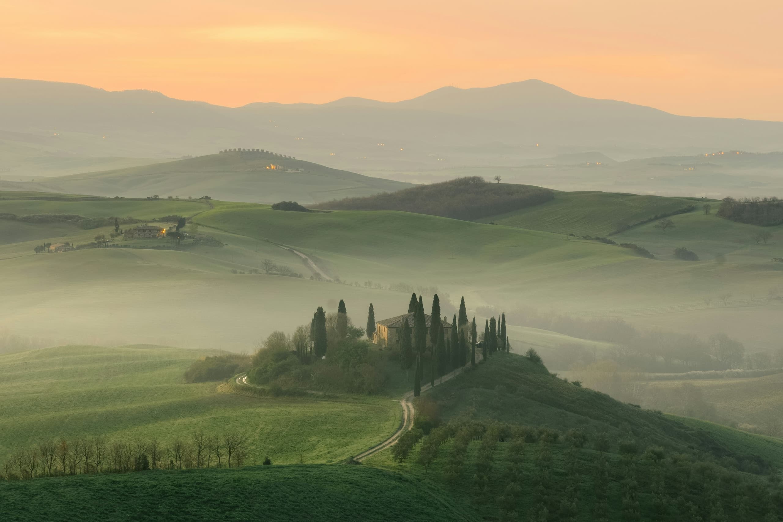 A lone farmhouse among rolling Tuscan hills glowing beneath a dramatic sunset